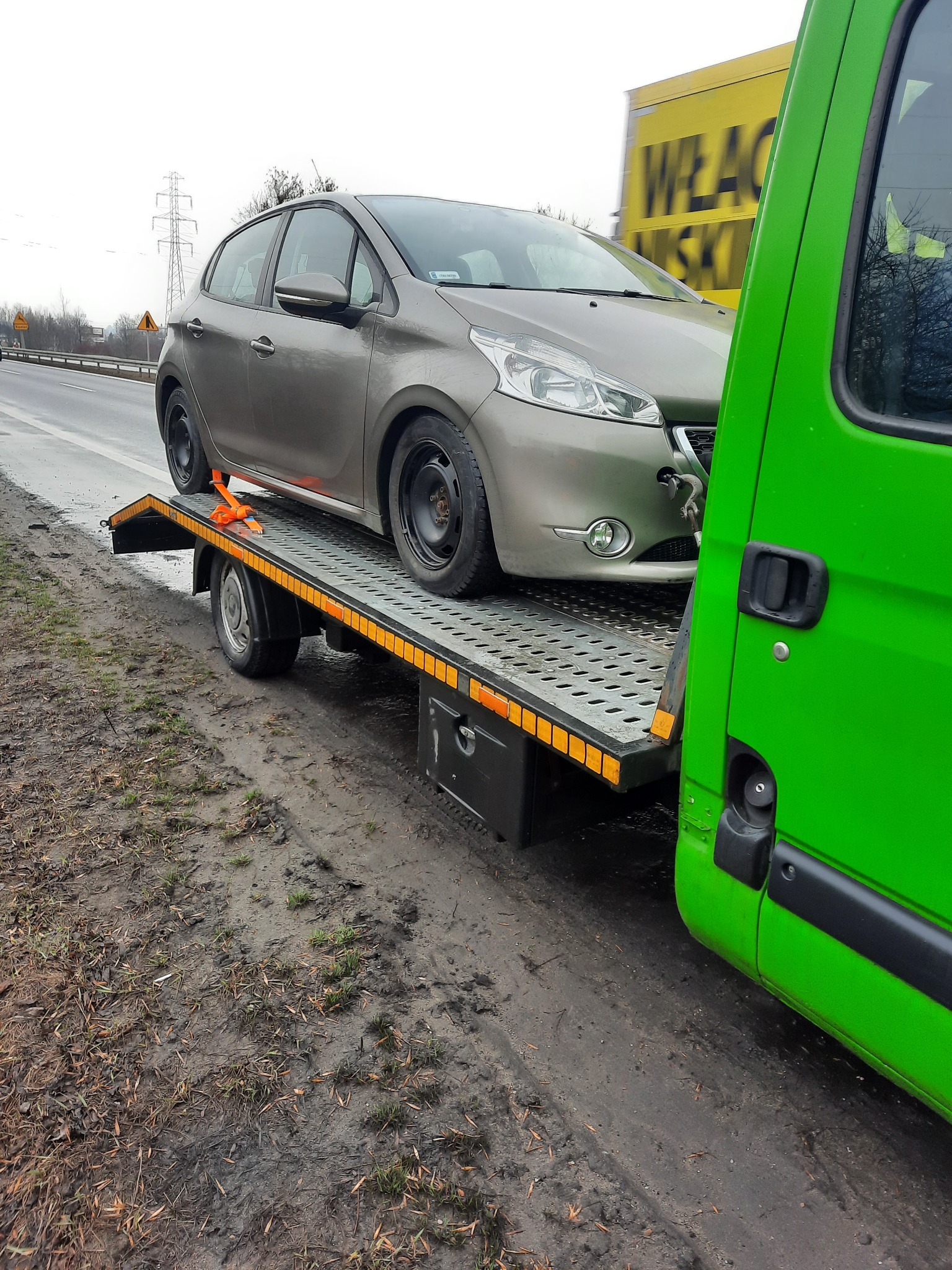 Szary samochód osobowy zabezpieczony pomarańczowymi pasami na platformie lawety, widok z boku, z zielonym bokiem pojazdu lawety po prawej stronie, w tle żółty billboard.