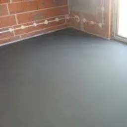 Gładka, ciemnoszara powierzchnia podłogi z widocznym fragmentem ściany z czerwonej cegły, na której zamontowane są białe rurki instalacyjne. Podłoga wydaje się być świeżo wylana i wykończona,...