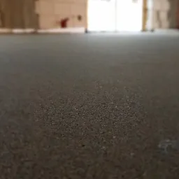 Świeżo wylana, szara posadzka w surowym stanie, z widocznymi bloczkami betonowymi w tle i otworami okiennymi w oddali.