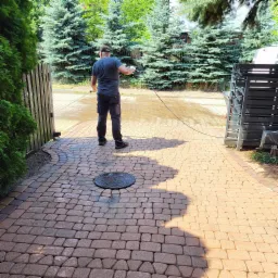 Mężczyzna w roboczych spodniach czyści kostkę brukową za pomocą myjki ciśnieniowej, widoczny manhole, w tle iglaki i brama.