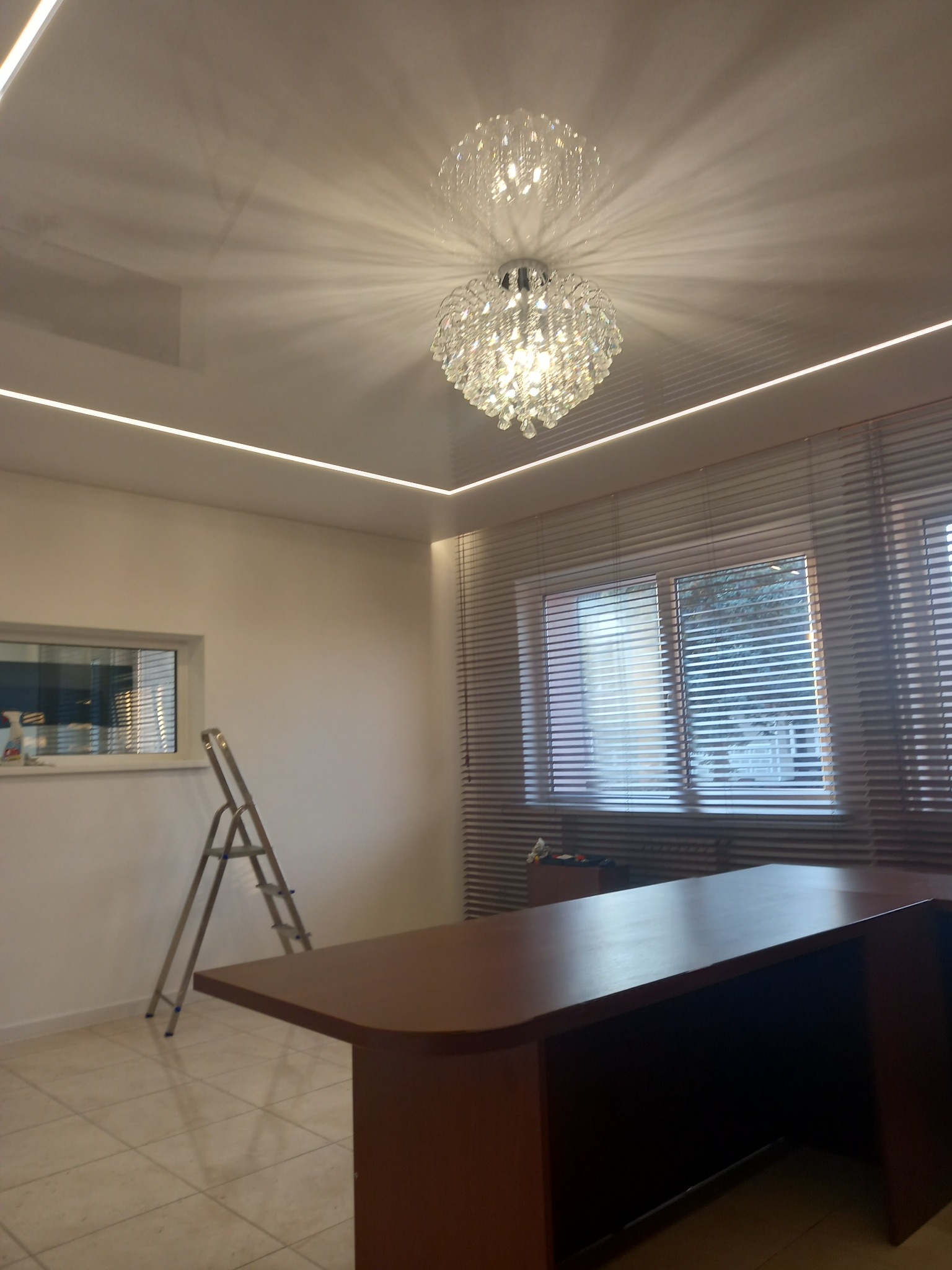 Kryształowy żyrandol odbijający światło na błyszczącym suficie z ledowym oświetleniem, drabina oparta o ścianę, brązowe biurko i okno z żaluzjami w tle.