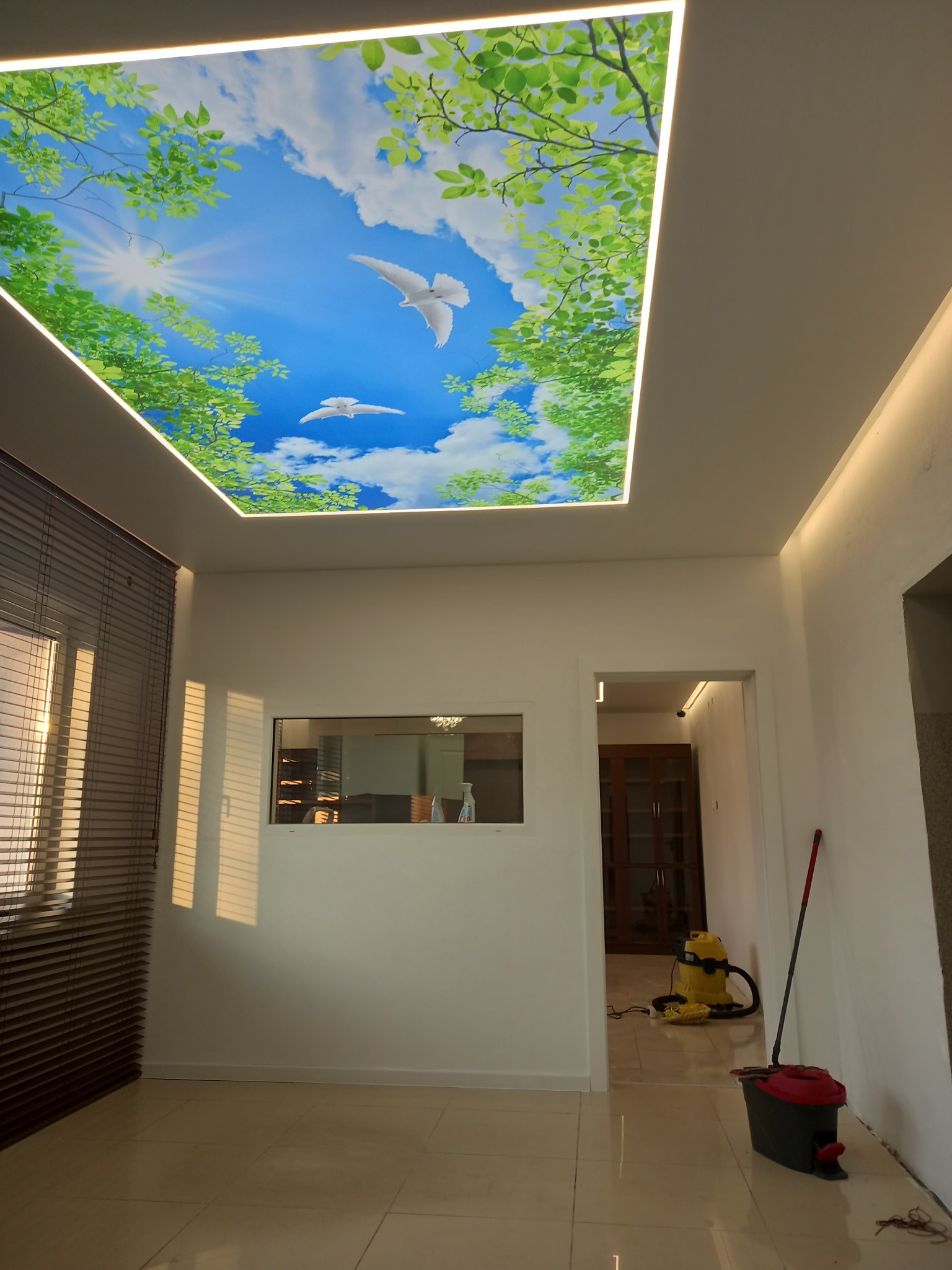 Pokój z podświetlanym sufitem napinanym z nadrukiem błękitnego nieba, białych chmur i zielonych liści, imitującym widok na zewnątrz.