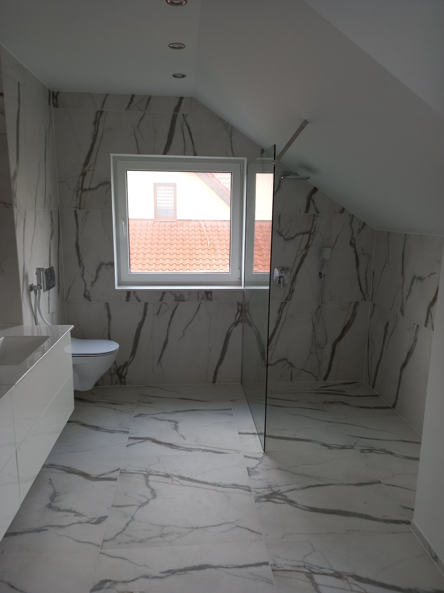 Nowoczesna łazienka na poddaszu z marmurowymi płytkami, kabiną prysznicową typu walk-in i oknem z widokiem na dachówkę.