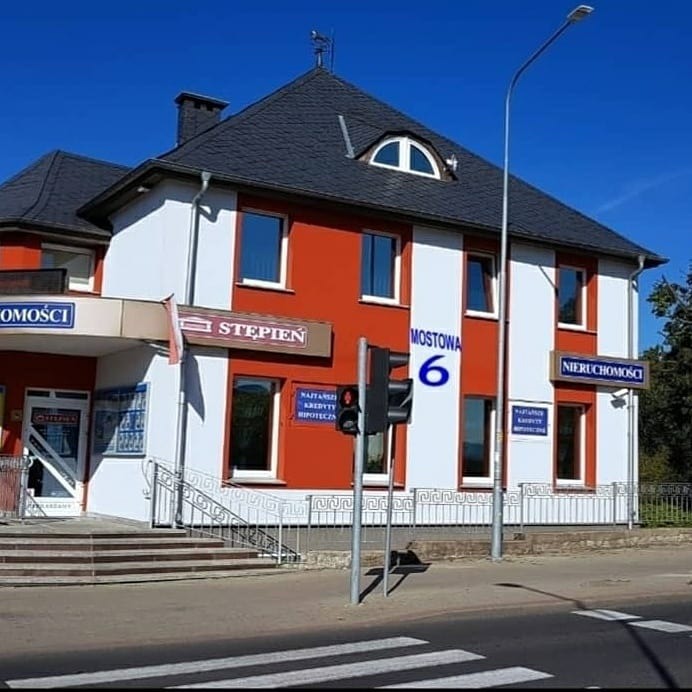 Dwupiętrowy budynek agencji nieruchomości z szyldami informacyjnymi i numerem 'Mostowa 6', z sygnalizacją świetlną w kadrze.