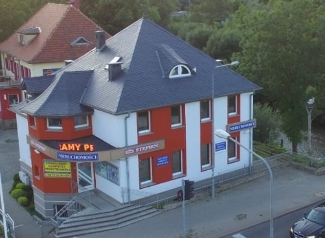 Dwupiętrowy budynek agencji nieruchomości z ciemnoszarym dachem, czerwonymi i białymi ścianami, oraz szyldami reklamowymi, widoczny z perspektywy ulicy.