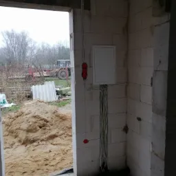 Wnętrze surowego budynku w trakcie instalacji elektrycznej: biała skrzynka rozdzielcza, kable prowadzone po ścianie z bloczków, widoczne puszki instalacyjne, na zewnątrz przez otwór okienny...