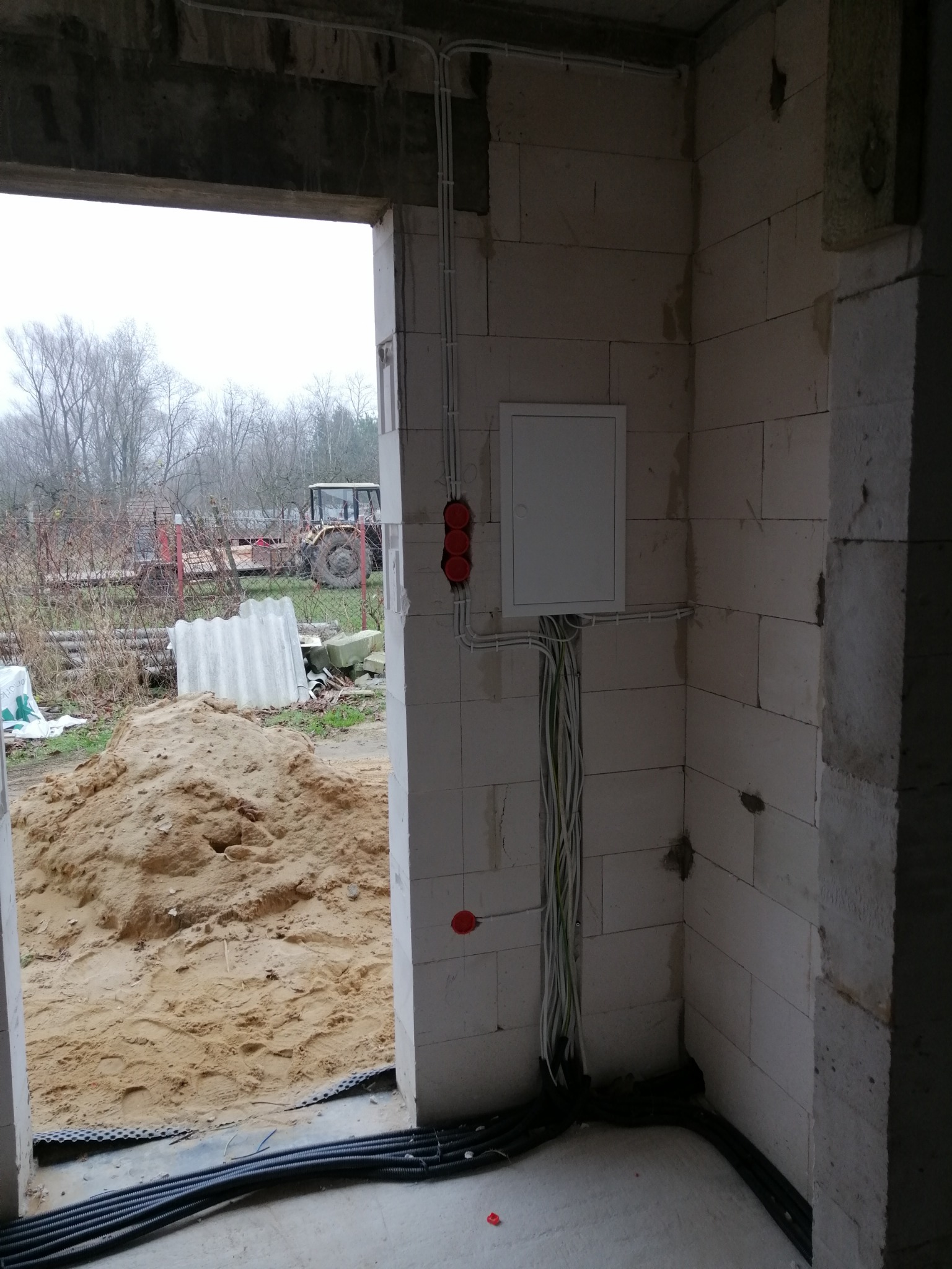 Wnętrze surowego budynku w trakcie instalacji elektrycznej: biała skrzynka rozdzielcza, kable prowadzone po ścianie z bloczków, widoczne puszki instalacyjne, na zewnątrz przez otwór okienny...