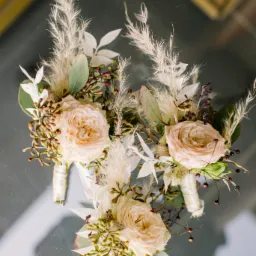 Trzy identyczne butonierki z kremowych róż, puszystych traw i zielonych liści na lustrzanym tle, każda owinięta białą wstążką.