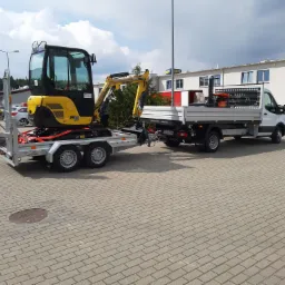 Żółta mini koparka Yanmar SV18 na przyczepie, ciągnięta przez biały samochód dostawczy z platformą, zaparkowane na kostce brukowej przed budynkiem Makro.