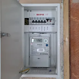 Otwarta skrzynka elektryczna z licznikiem i bezpiecznikami wewnątrz, widoczne przewody, zamontowana na ścianie w trakcie remontu, z widocznymi śladami po poprzednich instalacjach.