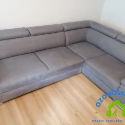 Szara, narożna kanapa tapicerowana z zagłówkami, stojąca na panelach podłogowych, z widocznym logo firmy oferującej pranie tapicerki.