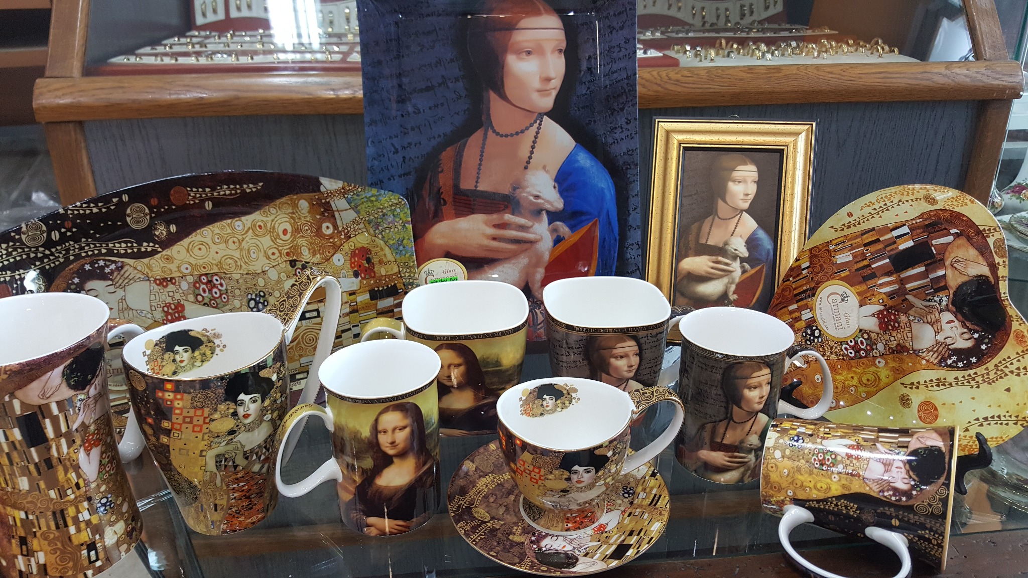 Zestaw upominkowy: filiżanki, talerzyki i tace z reprodukcjami obrazów Gustava Klimta i Leonarda da Vinci na szklanej półce w sklepie.