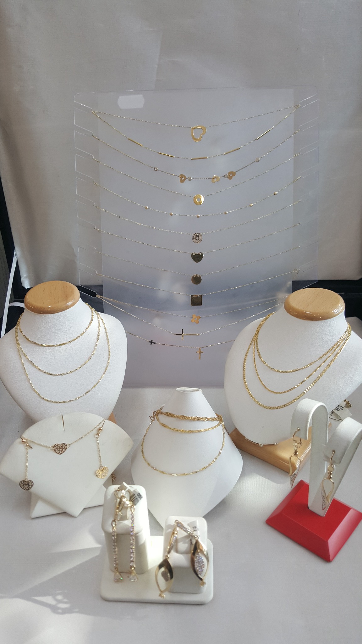 Złote naszyjniki i kolczyki prezentowane na białych ekspozytorach o różnych kształtach, część w przezroczystym organizerze z kilkoma poziomami, z zawieszkami w kształcie serc, kółek, krzyżyków...