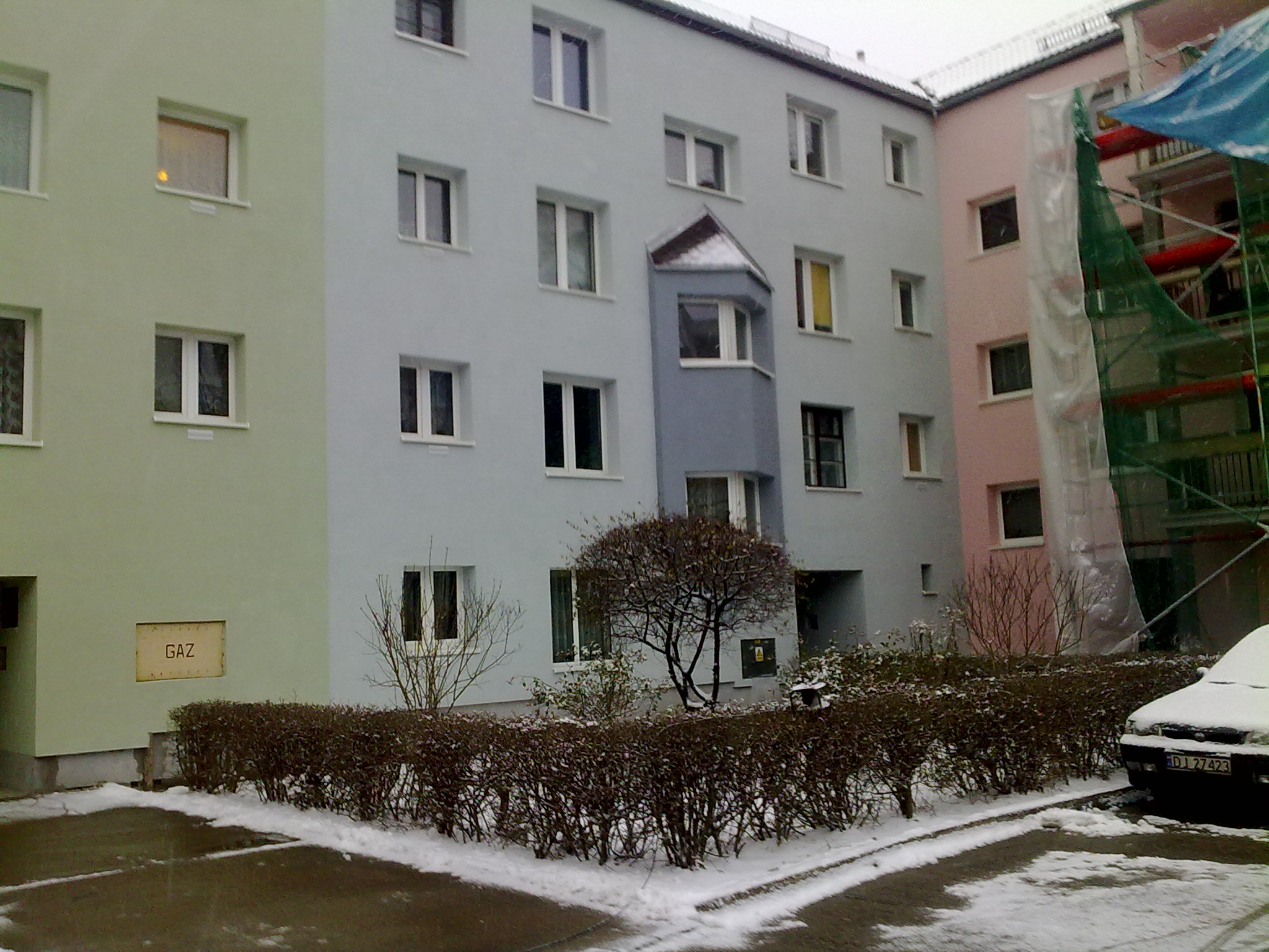 Wielobarwny budynek wielorodzinny z oknami, częściowo ośnieżony, z widocznymi rusztowaniami i siatką ochronną przy jednej ze ścian.