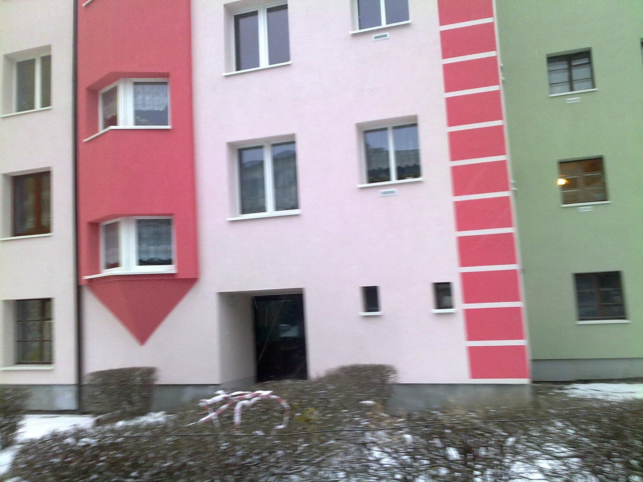 Wielokolorowa elewacja budynku wielorodzinnego z oknami i wejściem, częściowo pokryta śniegiem w krzewach.