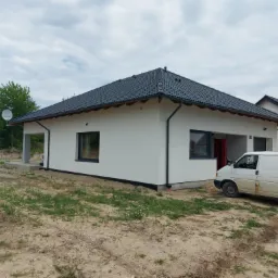 Nowoczesny parterowy dom z ciemną dachówką, białymi ścianami i szarą stolarką okienną, na działce z piaskiem i ruderalną roślinnością, z zaparkowanym białym busem obok wejścia.