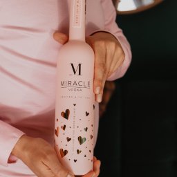 WIELKOPOLSKA MANUFAKTURA WÓDEK SPÓŁKA Z OGRANICZONĄ ODPOWIEDZIALNOŚCIĄ - Różowa butelka wódki Miracle z serduszkami, trzymana przez osobę w różowej koszuli, na tle zielonej ściany i lustra. Edycja limitowana Rose Gold.