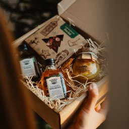 WIELKOPOLSKA MANUFAKTURA WÓDEK SPÓŁKA Z OGRANICZONĄ ODPOWIEDZIALNOŚCIĄ - Otwarty kartonowy kosz prezentowy z nalewkami w małych butelkach i słoikiem cytryn z imbirem, wypełniony siankiem, trzymany w dłoni.