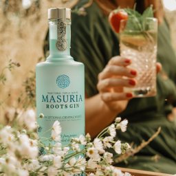 WIELKOPOLSKA MANUFAKTURA WÓDEK SPÓŁKA Z OGRANICZONĄ ODPOWIEDZIALNOŚCIĄ - Butelka ginu Masuria Roots Gin z koktajlem na drewnianej tacy, wśród polnych kwiatów. Kobieta w tle trzyma drinka, tworząc letnią, relaksującą atmosferę.