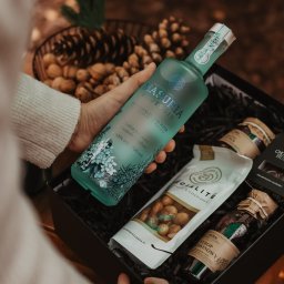 WIELKOPOLSKA MANUFAKTURA WÓDEK SPÓŁKA Z OGRANICZONĄ ODPOWIEDZIALNOŚCIĄ - Kosz prezentowy: ręka trzyma butelkę ginu Masuria Roots Gin, obok orzechy w czekoladzie, syrop i czekoladki. W tle szyszki i orzechy laskowe.