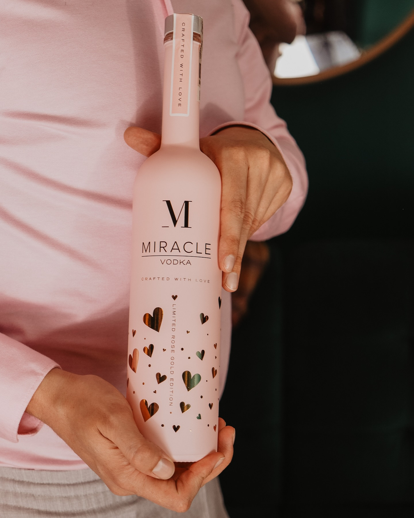 Różowa butelka wódki Miracle z serduszkami, trzymana przez osobę w różowej koszuli, na tle zielonej ściany i lustra. Edycja limitowana Rose Gold.