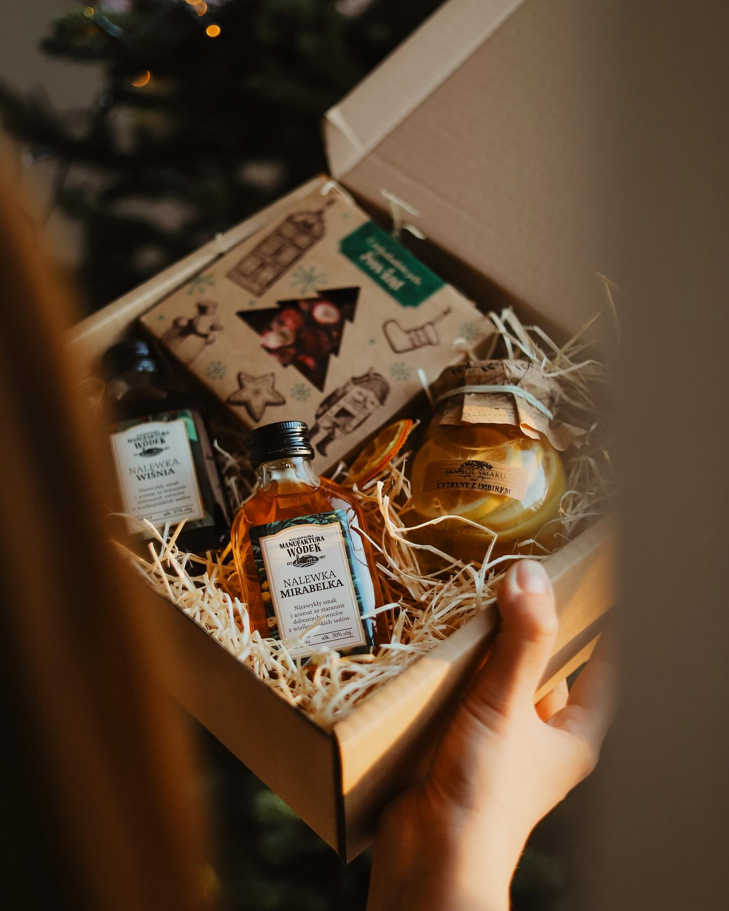 Otwarty kartonowy kosz prezentowy z nalewkami w małych butelkach i słoikiem cytryn z imbirem, wypełniony siankiem, trzymany w dłoni.