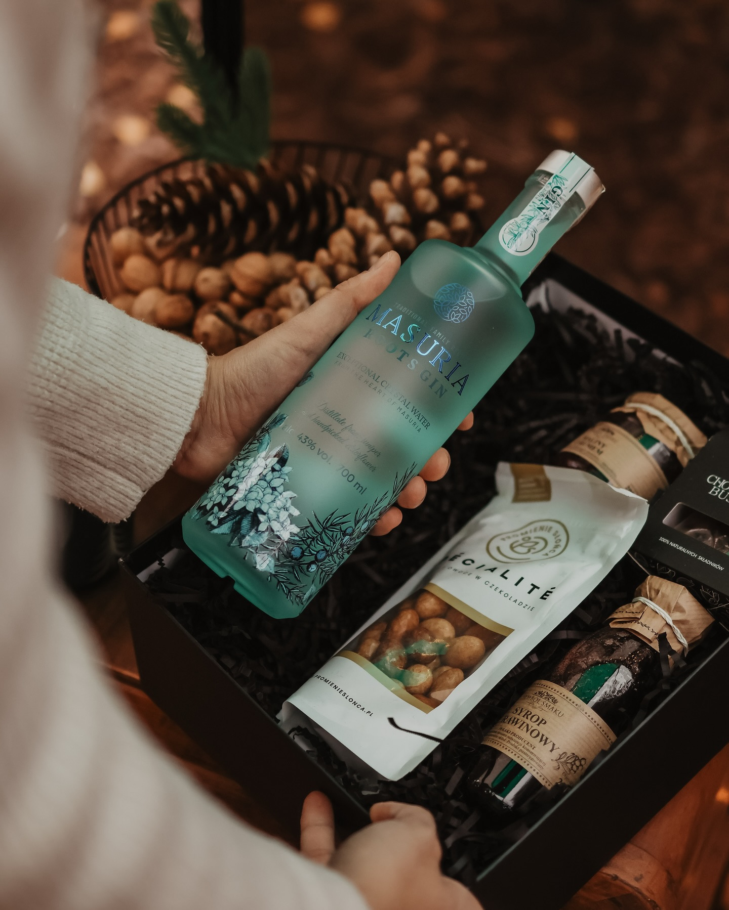 Kosz prezentowy: ręka trzyma butelkę ginu Masuria Roots Gin, obok orzechy w czekoladzie, syrop i czekoladki. W tle szyszki i orzechy laskowe.
