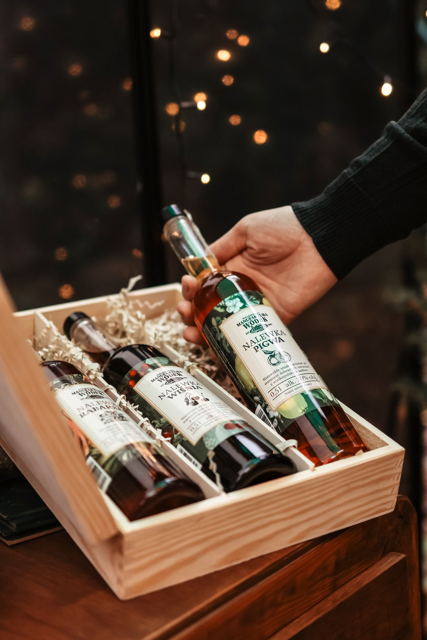 Drewniana skrzynka z trzema butelkami nalewek Manufaktura Wódek, trzymana w dłoni na tle migoczących światełek. Nalewki: Pigwa, Wiśnia, Rabarbar.