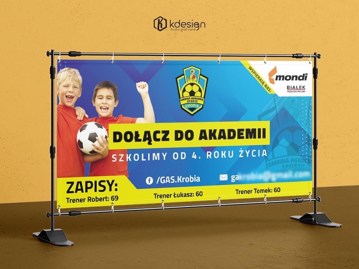 Baner reklamowy szkoły sportowej z uśmiechniętymi chłopcami trzymającymi piłkę, na tle żółtej ściany. Widoczne logo szkoły, hasło 'Dołącz do Akademii' i informacje o zapisach oraz trenerach.