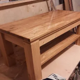 Stolik kawowy z jasnego drewna dębowego z dolną półką, widoczny w warsztacie stolarskim, wykończony lakierem o wysokim połysku.
