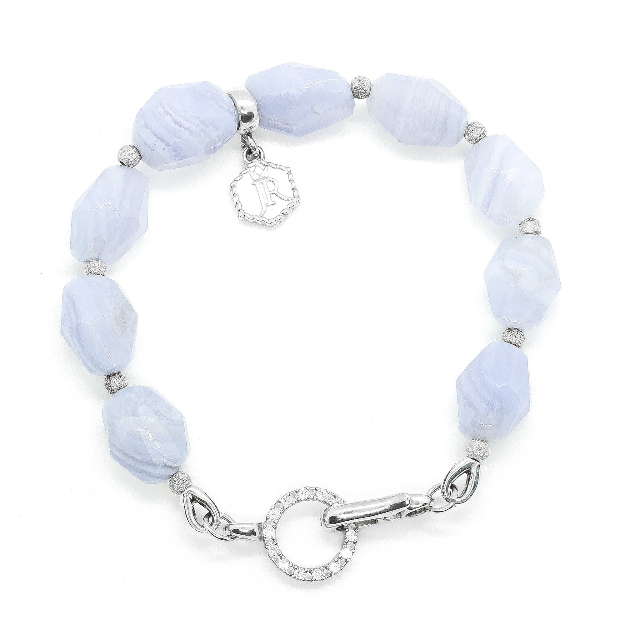 Bransoletka z fasetowanych, błękitnych kamieni i srebrnych kuleczek z cyrkoniami, z zawieszką z monogramem JR i zapięciem w kształcie pierścienia wysadzanego cyrkoniami, na białym tle.