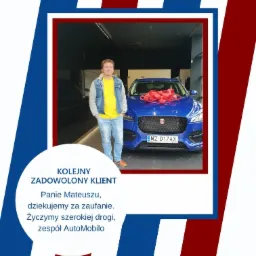 Mężczyzna w jeansowej kurtce i ż&oacute;łtej koszulce pozuje obok niebieskiego samochodu z czerwoną kokardą na masce, w salonie samochodowym.