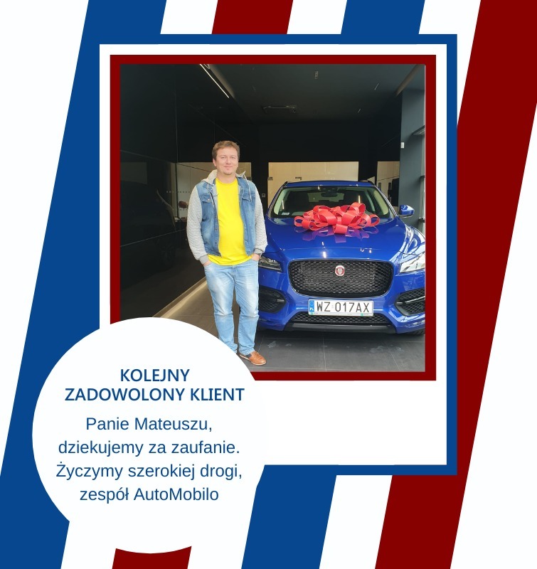 Mężczyzna w jeansowej kurtce i żółtej koszulce pozuje obok niebieskiego samochodu z czerwoną kokardą na masce, w salonie samochodowym.