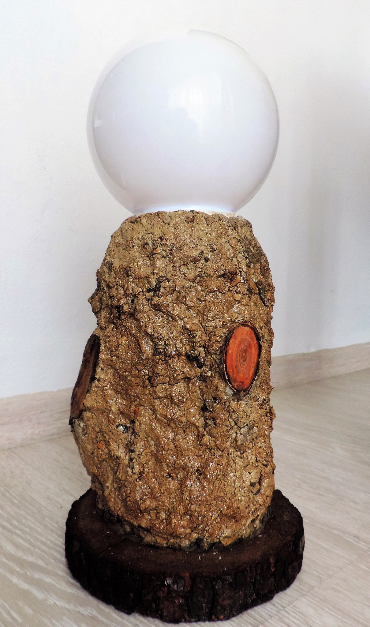 Lampa stołowa z podstawą z kory drzewnej i kulistym, białym kloszem na jasnej podłodze.