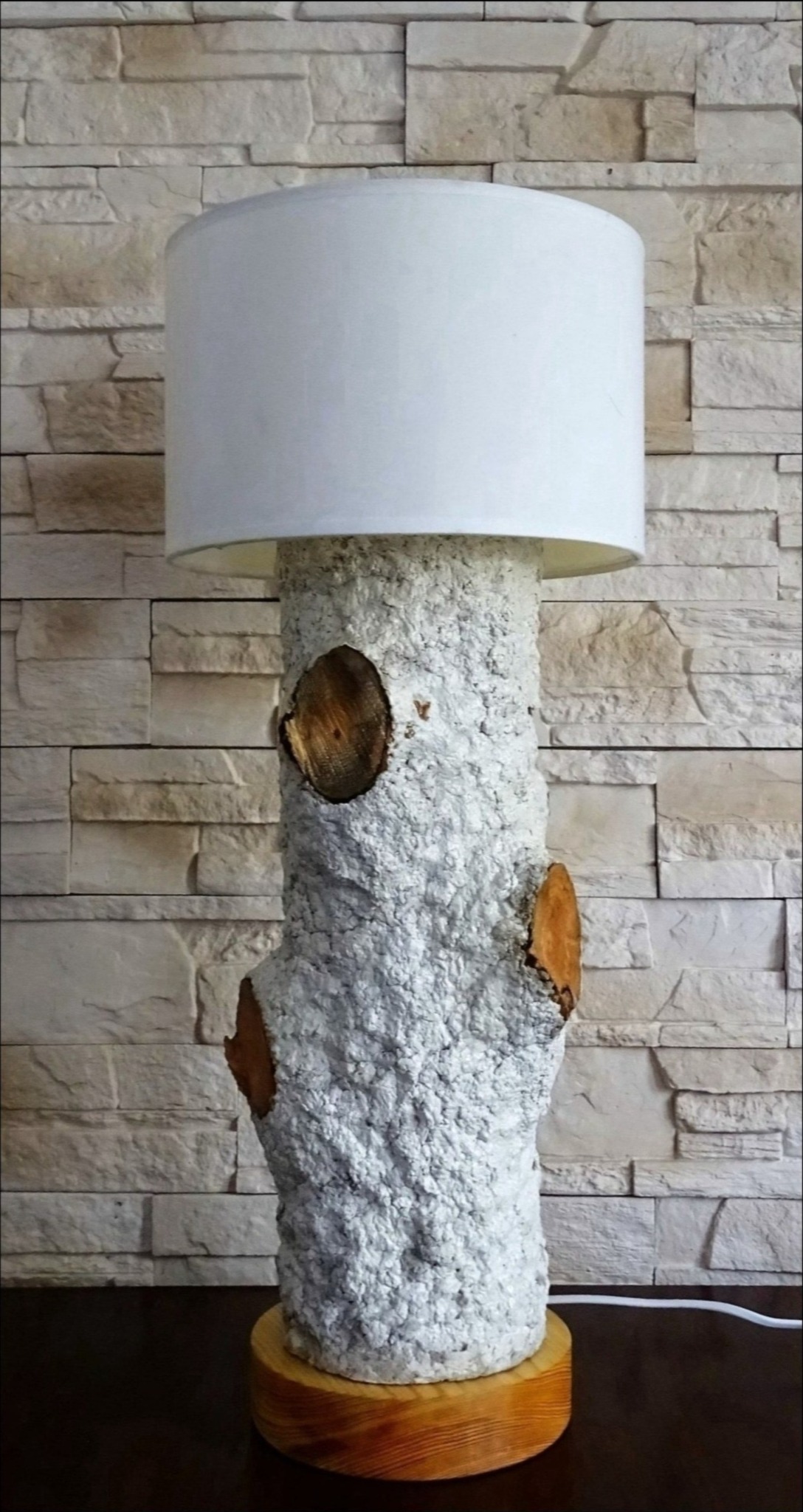Lampa stojąca z abażurem, wykonana z pnia drzewa z widocznymi sękami, umieszczona na drewnianej podstawie, na tle ściany z kamienia dekoracyjnego.