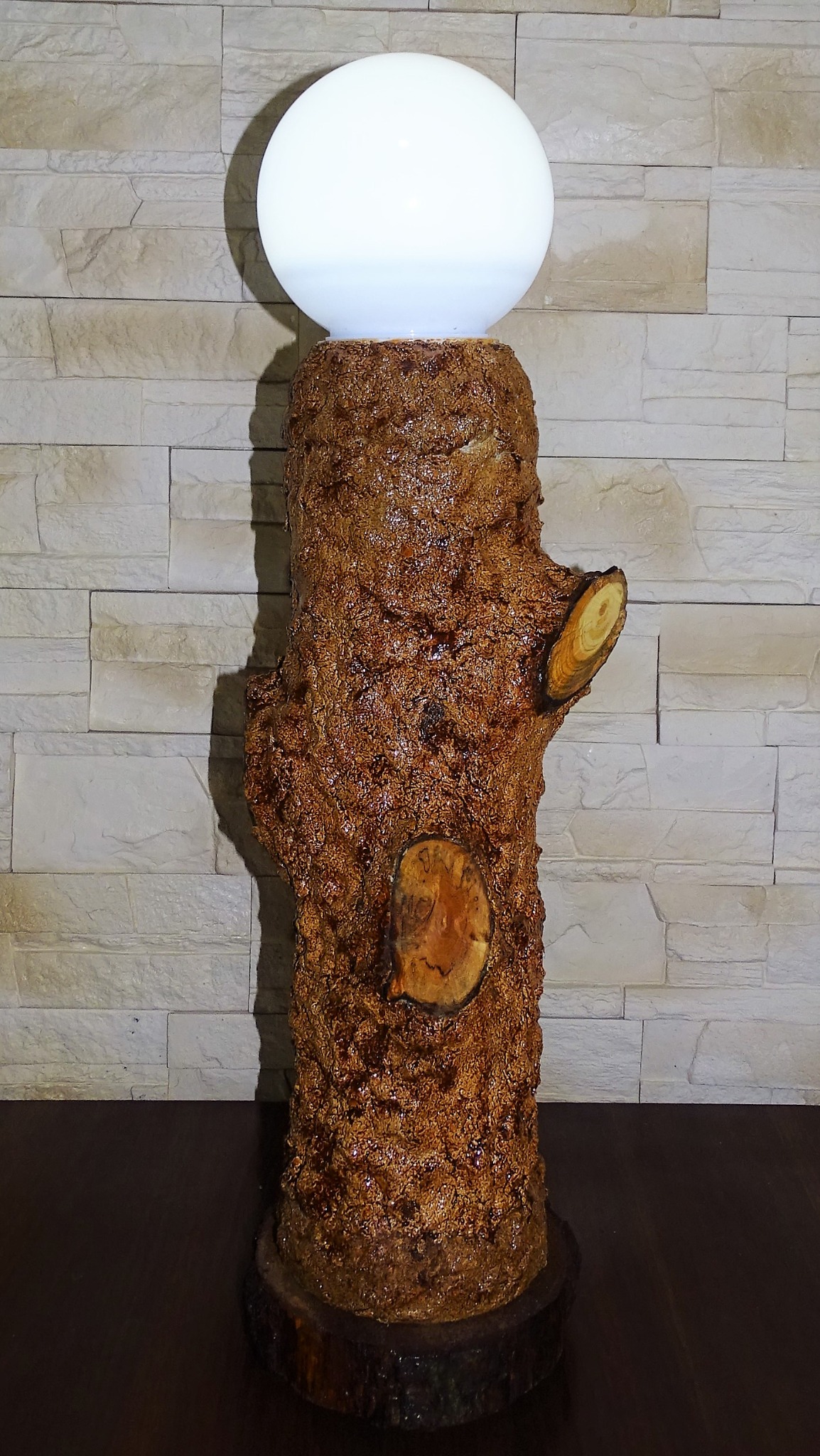Lampa stojąca z drewnianego pnia z okrągłym, białym kloszem, widoczne sęki i faktura drewna, tło z cegieł.