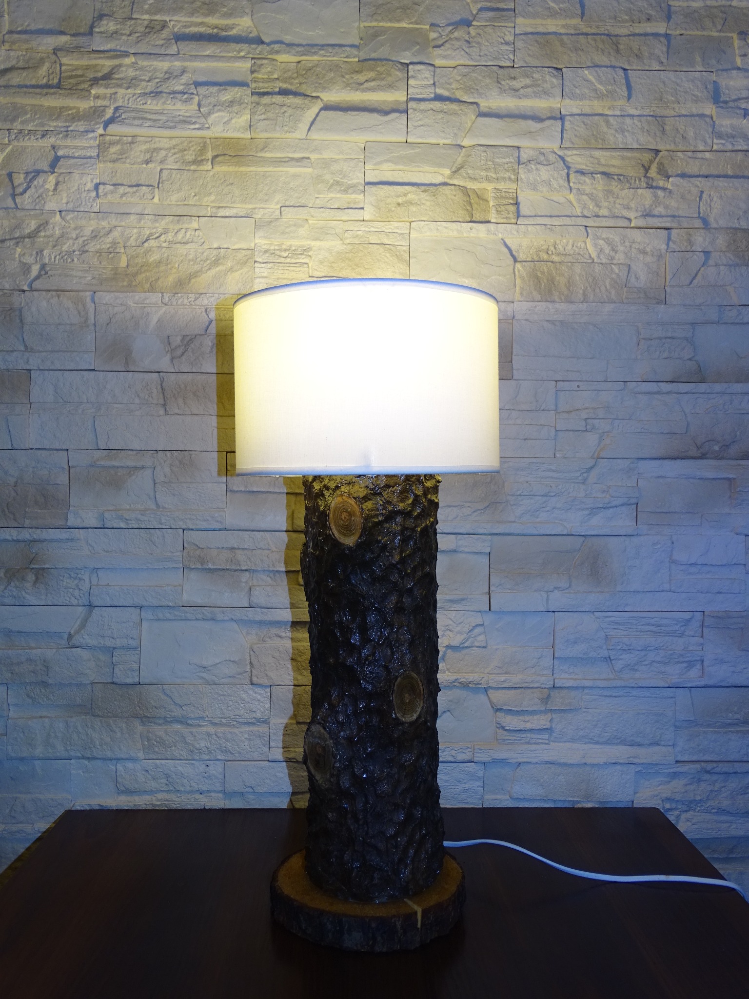 Lampa stołowa z podstawą z naturalnego drewna, z widocznymi słojami i białym abażurem, na tle ściany z dekoracyjnej cegły.
