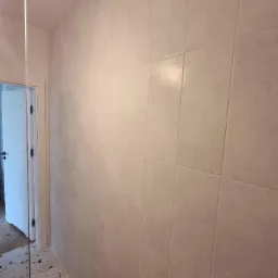 Wykończona ściana w łazience z jasnymi, kwadratowymi płytkami ceramicznymi, widoczny fragment podłogi lastryko i rama szklanej kabiny prysznicowej oraz wystający element instalacji hydraulicznej.