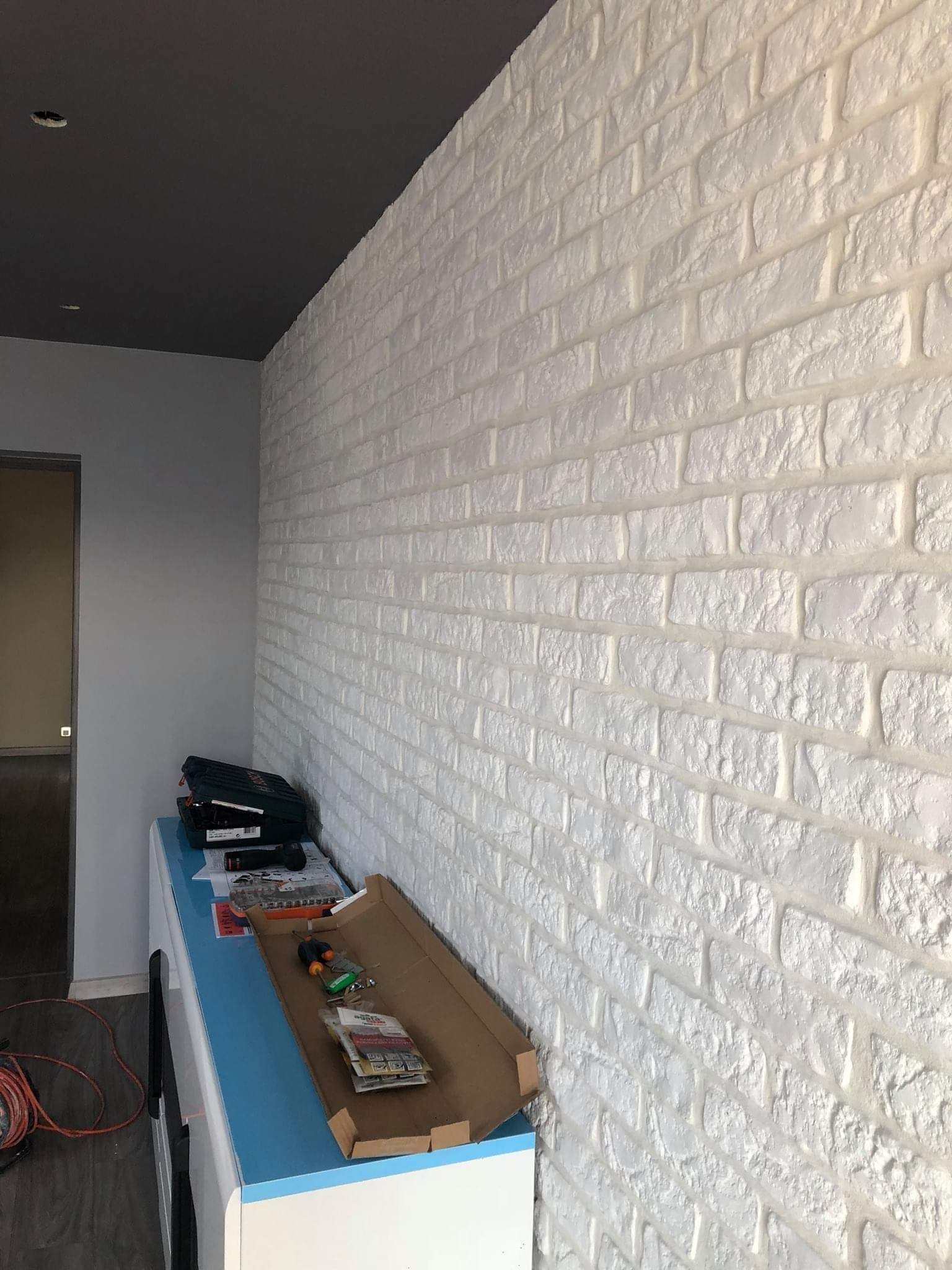 Ściana wykończona białymi panelami imitującymi cegłę, z widocznymi narzędziami i akcesoriami na szafce w tle, sufit pomalowany na ciemny szary kolor.