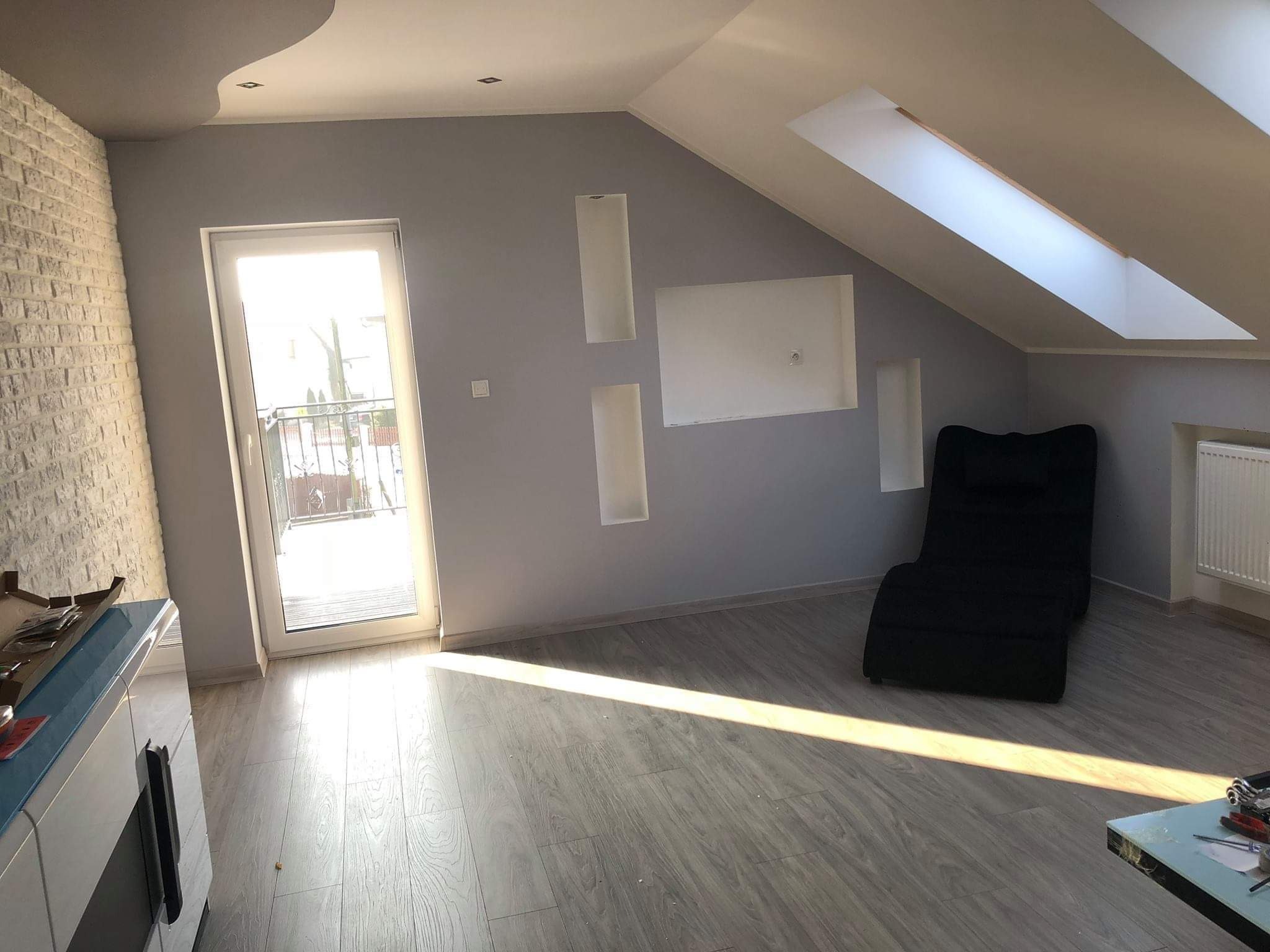 Poddasze po remoncie z szarą ścianą, imitacją cegły po lewej, wnękami w ścianie, oknem dachowym i jasną podłogą z paneli.