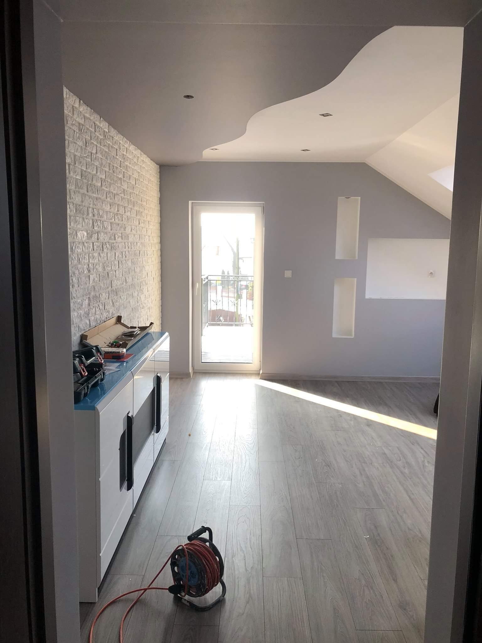 Wnętrze poddasza po remoncie, z białą cegłą na ścianie, szarą farbą na pozostałych ścianach, drewnianą podłogą i dekoracyjnym sufitem podwieszanym. Widoczne narzędzia na komodzie i zwinięty...