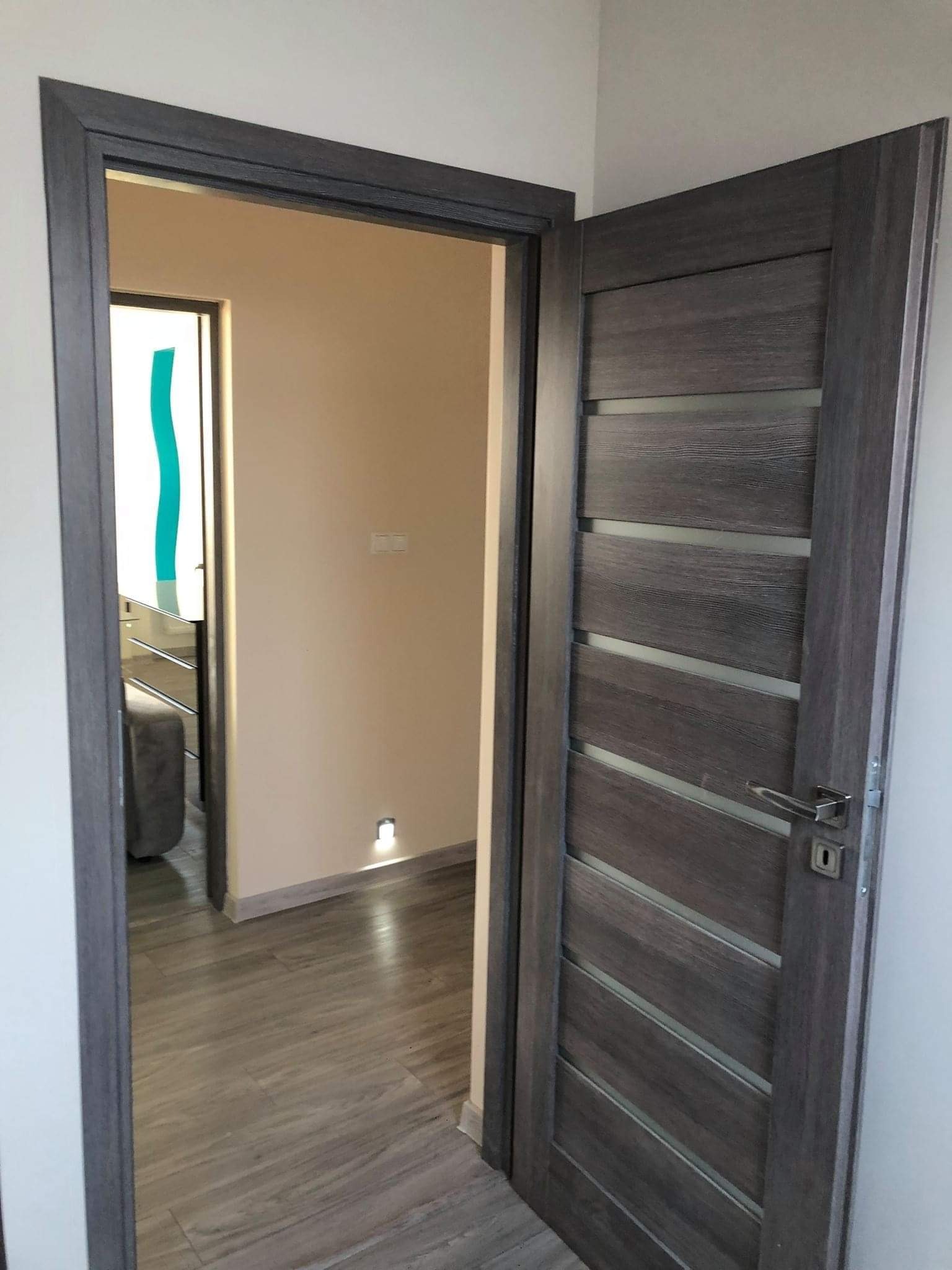 Otwarta, nowoczesna, szara, drewnopodobna wewnętrzna stolarka drzwiowa z przeszkleniami, widoczna na tle jasnego wnętrza z drewnianą podłogą i delikatnym oświetleniem podłogowym.