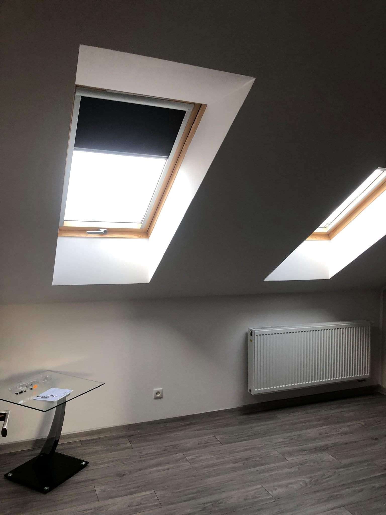 Poddasze z dwoma oknami dachowymi, jedno z roletą, grzejnik na ścianie, szare panele podłogowe i stolik ze szklanym blatem.
