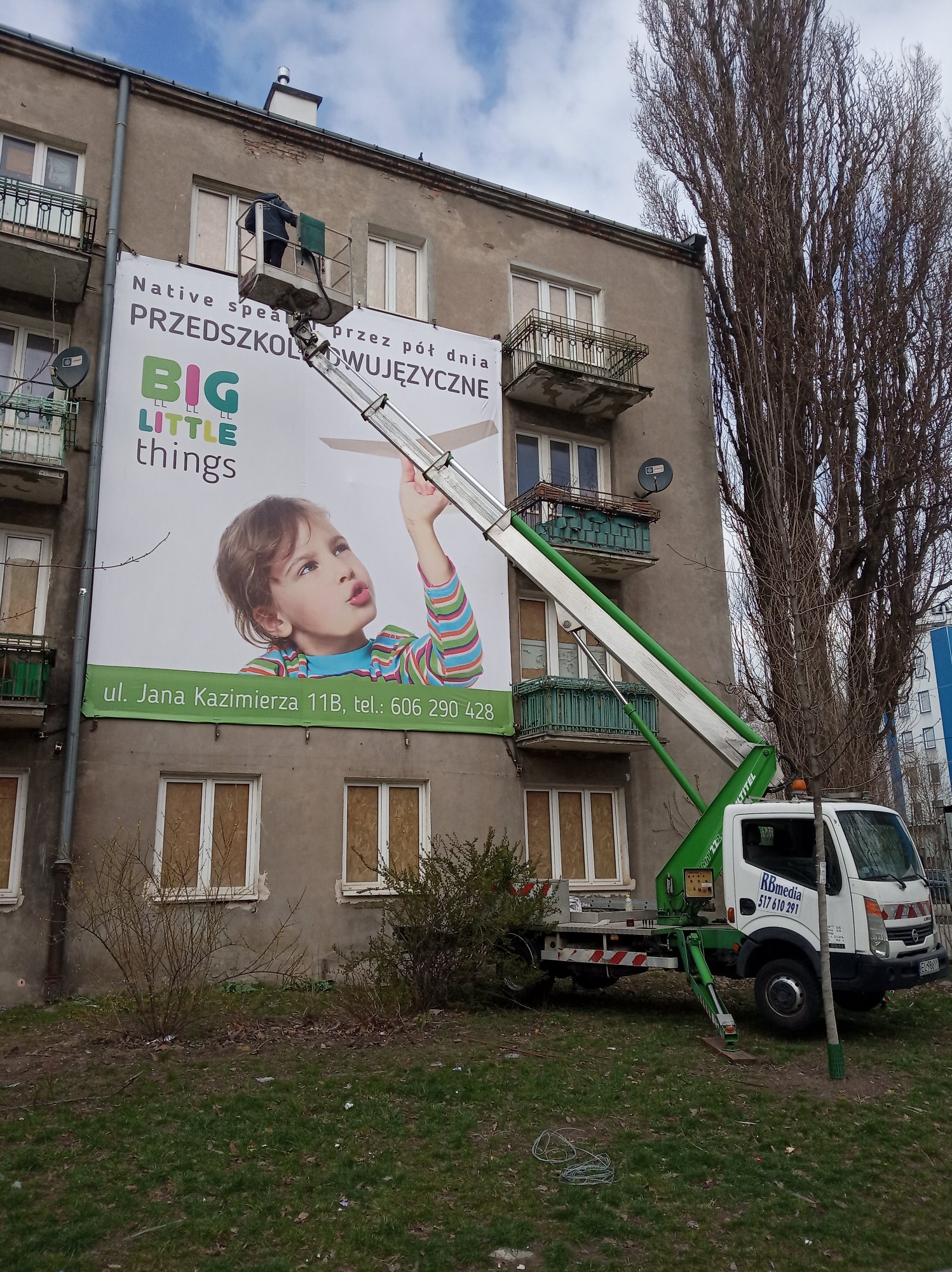 Montaż wielkoformatowej reklamy przedszkola językowego na elewacji budynku mieszkalnego z użyciem podnośnika koszowego, widoczne okna zabite płytami.
