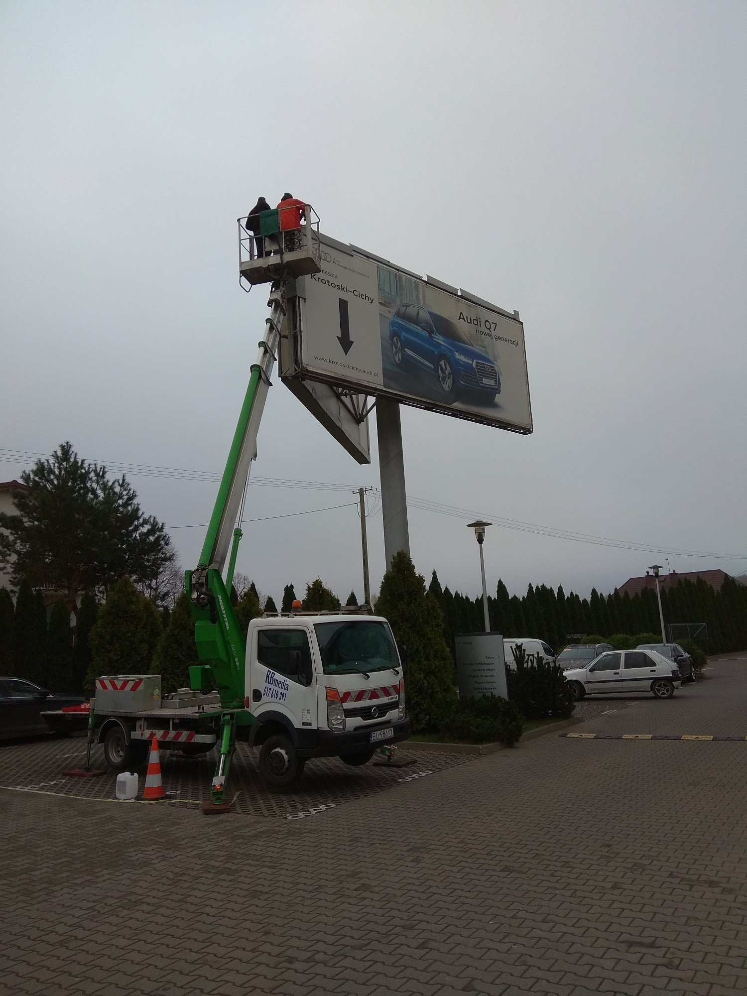 Montaż billboardu reklamowego Audi Q7 z wykorzystaniem podnośnika koszowego na parkingu, widoczne dwie osoby w koszu oraz zaparkowane samochody w tle.