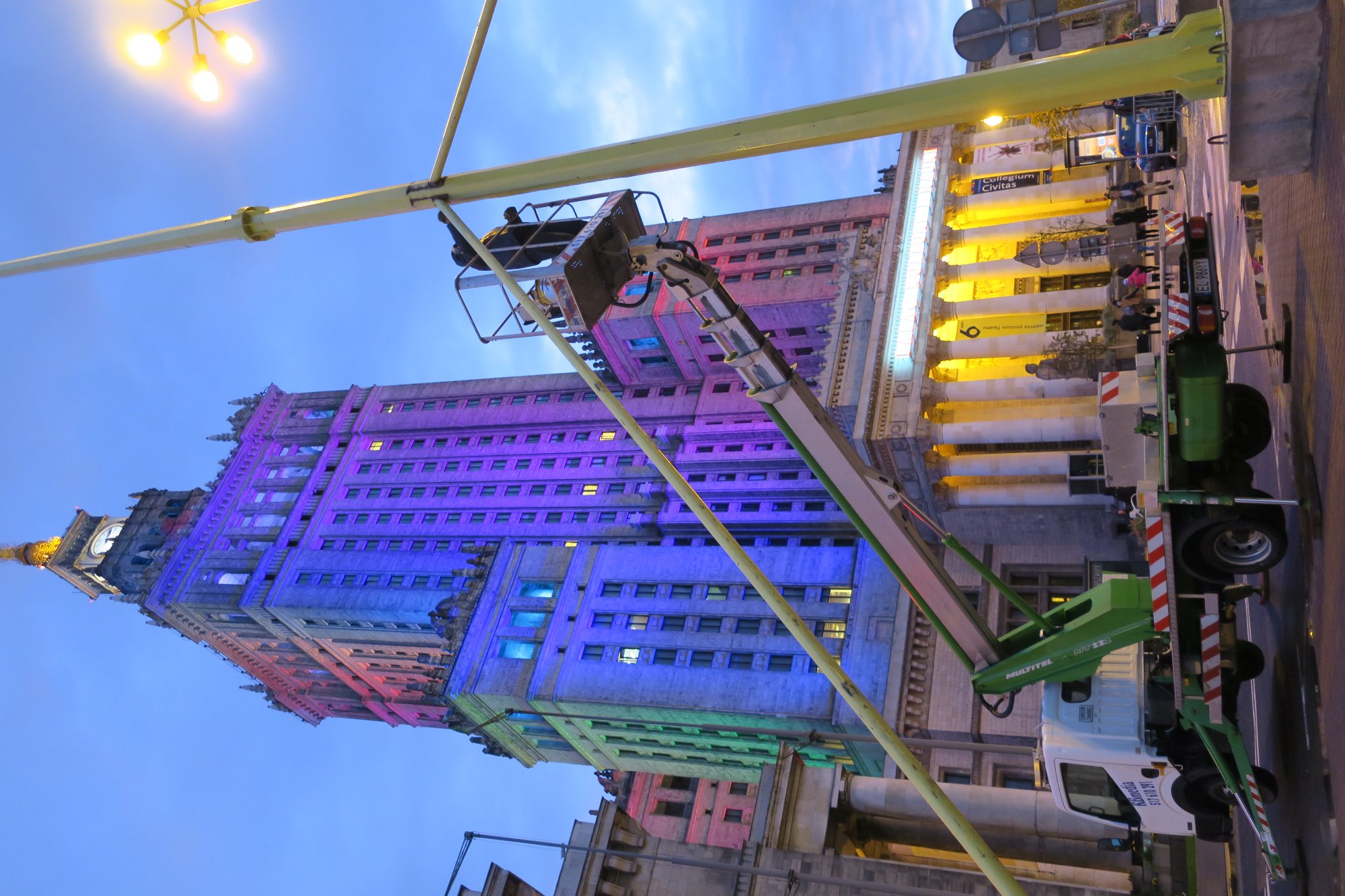 Wysięgnik koszowy na tle podświetlonego kolorami Pałacu Kultury w Warszawie, widok z dołu, wieczorne oświetlenie.