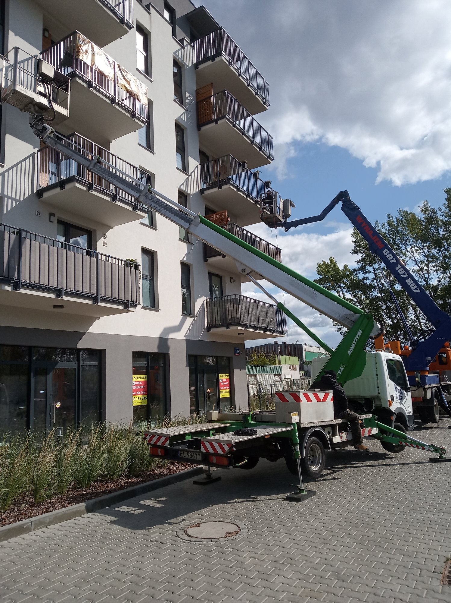 Dwie podnośniki koszowe ustawione przy nowoczesnym budynku mieszkalnym z balkonami, jeden z koszem na wysokości balkonu, prawdopodobnie podczas prac montażowych lub transportowych.