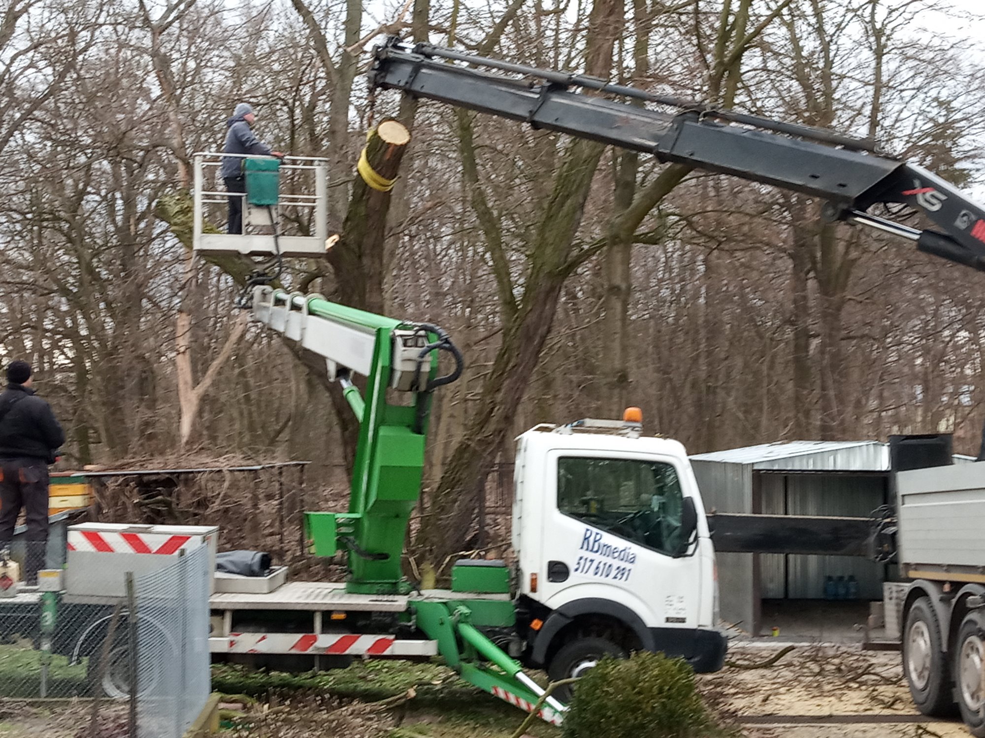 Pracownik w podnośniku koszowym przycina drzewo na wysokości, widoczny dźwig i samochód z logo firmy na tle drzew bez liści.