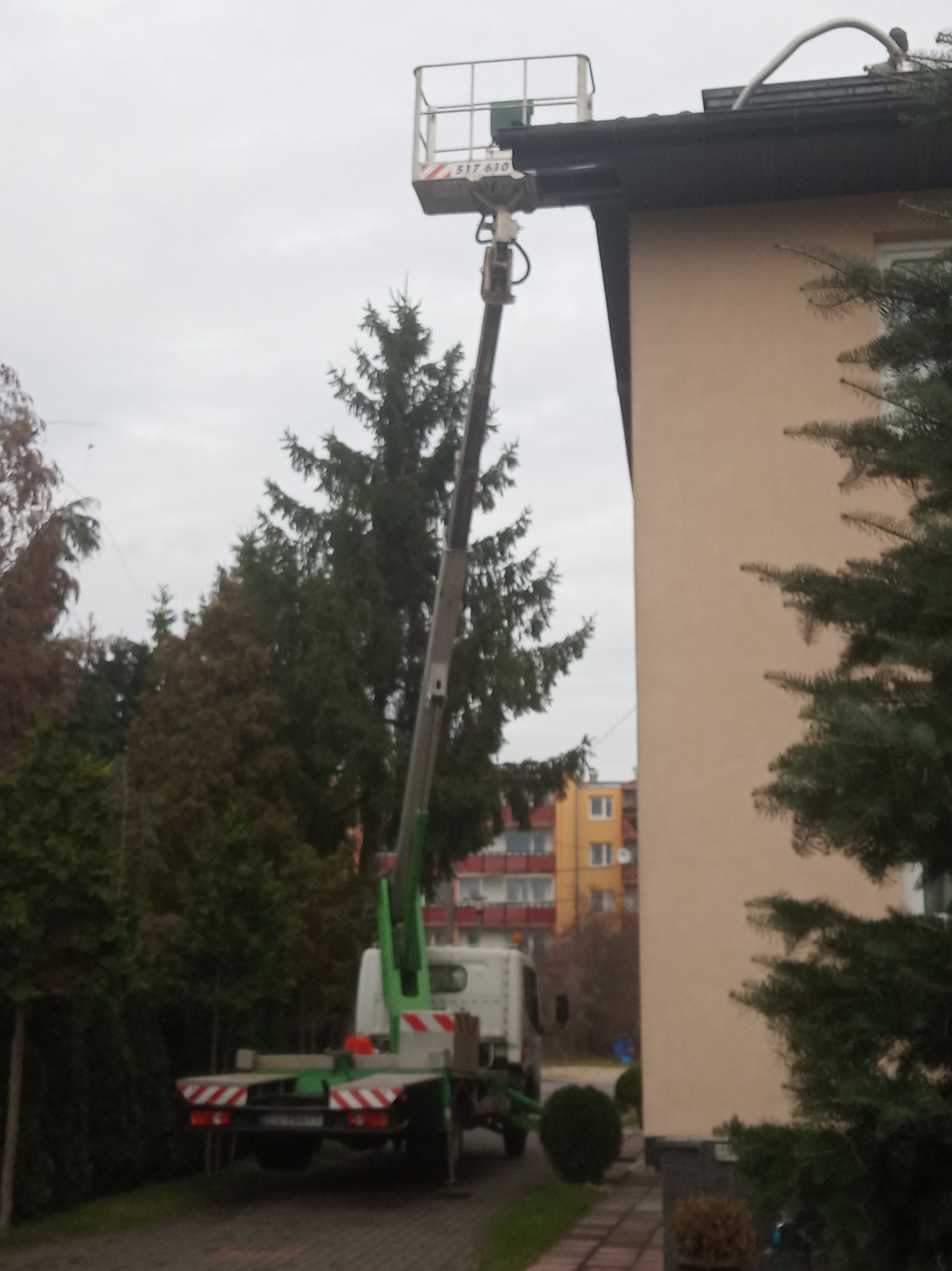 Podnośnik koszowy ustawiony przy ścianie budynku, prawdopodobnie podczas prac konserwacyjnych dachu. Widoczna platforma z osobą oraz przewód wchodzący na dach. W tle drzewa i budynek mieszkalny.