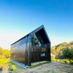 Nowoczesny dom szkieletowy z ciemnego drewna z dużym oknem szczytowym, otoczony zielenią i błękitnym niebem.